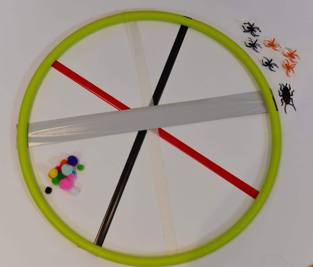 spider web hula hoop for a science activity. Four different types of sticky  tape are stretched across the hula hoop. coloured pom poms and plastic spiders sit to the side.