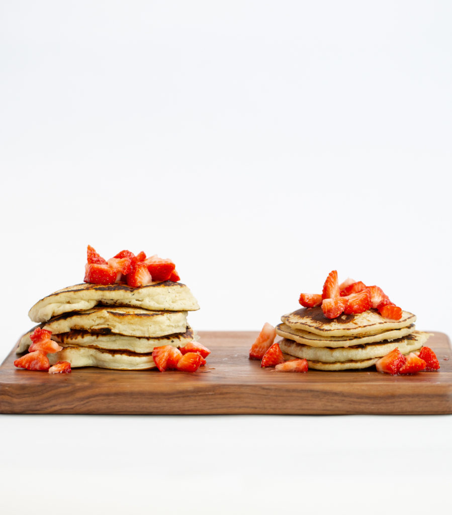 two piles of pancakes with strawberries and syrup on top. One pile is big and fluffy the other smaller and thinner