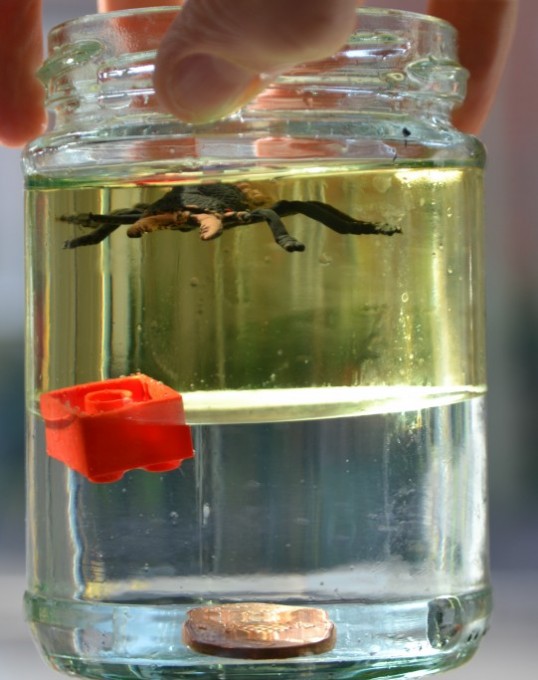 Density jar made with oil and water. a coin sits on the bottom, a lego brick between the oil and water and a plastic spider on the top