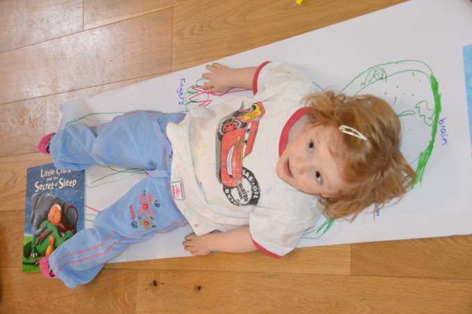young girl lying on a large long sheet of paper. The girls outline has been drawn around her in felt tip