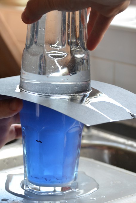 Two glasses filled with liquid. The bottom one contains blue food colouring, water and salt. The top glass is plain water. The water glass is on top of the blue water glass with a piece of card in between them.