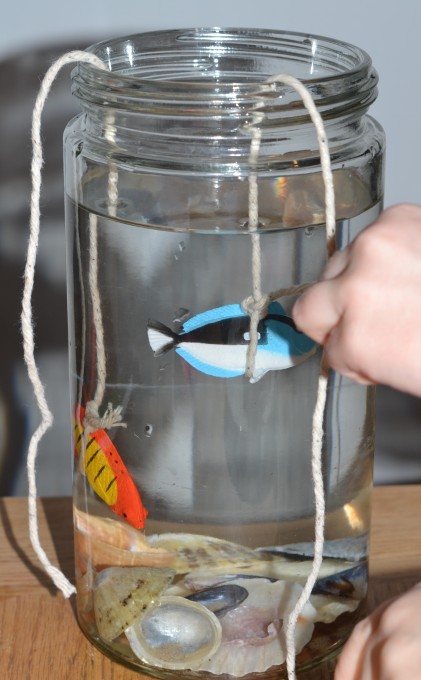 Jar of water with sand at the bottom and toy fish. The jar has litter and black food colouring in to demonstrate the effect of pollution of marine animals.