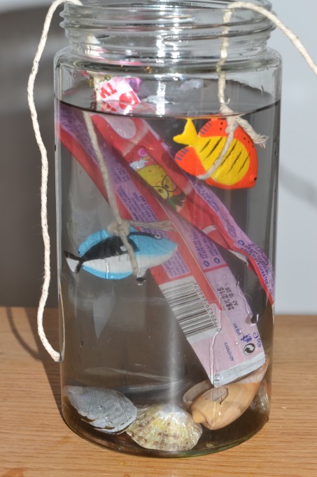 Glass jar with plastic fish and shells, the clear water has been made murky with the addition of black food colouring and plastic rubbish has been added too.