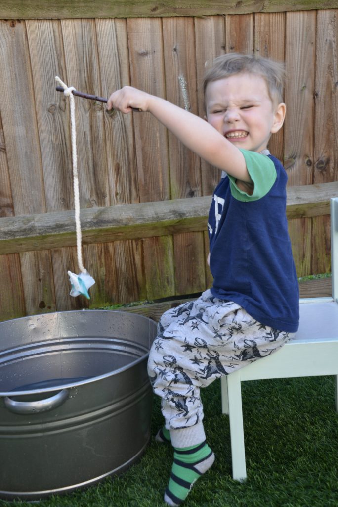 small boy sat on a wooden chair outdoors holding a stick with a piece of string with an ice cup attached to it