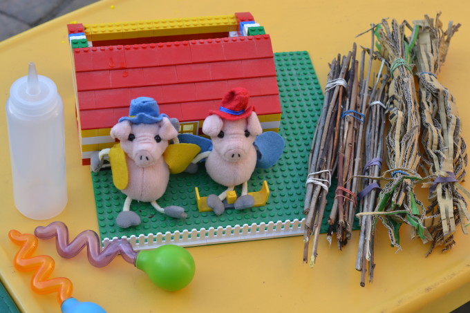 Image of three soft pig puppets sat outside a LEGO house with sticks and droppers at the side