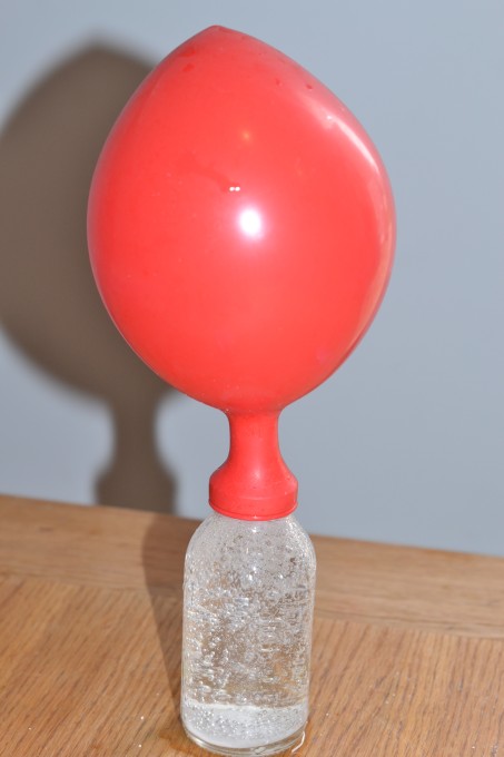 blow up a balloon with alka seltzer science activity. Image shows a red balloon on top of a small jar containing water and alka seltzer