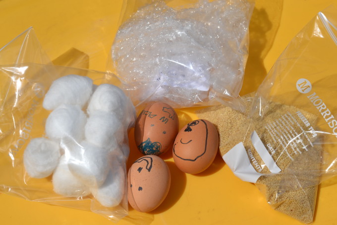 Eggs with faces drawn on them next to three plastic transparent bags. One is filled with cotton balls, one bubble wrap and one cous cous