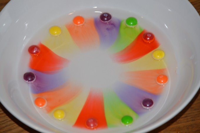 Skittles arranged in a circle on a white plate. Water has been poured over the top and colour from the skittles has diffused into the water.