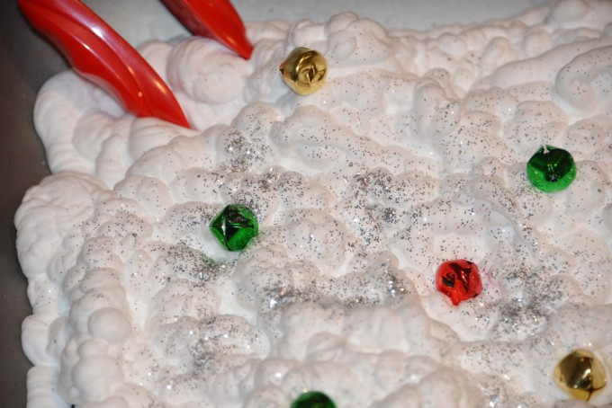 Christmas fine motor skill idea. Help the elves find their lost jingle bells! Image shows small coloured bells in a tray of shaving cream with glitter sprinkled on the top