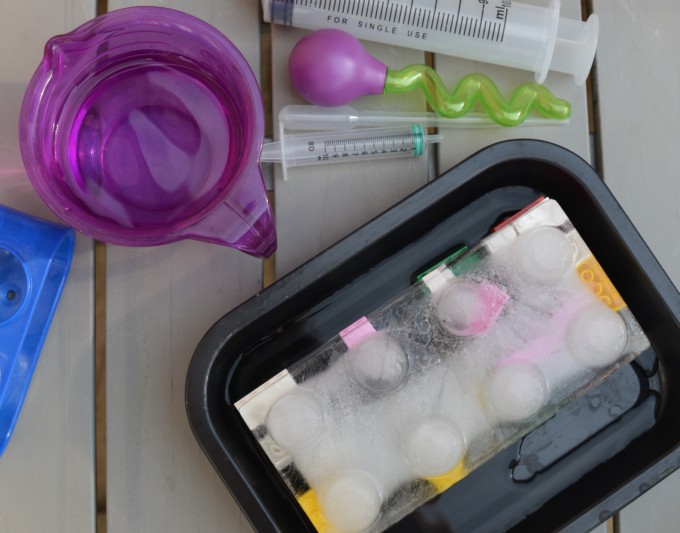 large block of ice in a black tray with lego bricks frozen inside. There are also syringes, droppers and a purple container on the table