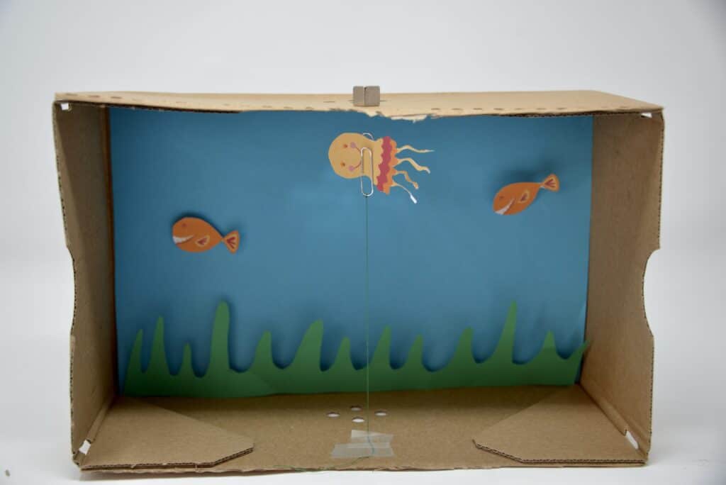 a cardstock octopus seeming to float in a shoebox. The octopus is attached to a paperclip and length of string. A magnet on top of the box is attracted to the paperclip making it look like the octopus is floating