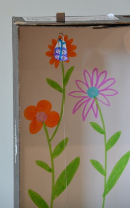 A flower drawing with a paperclip attached. The flower is floating in the air as it is attracted to a magnet on top of a shoebox