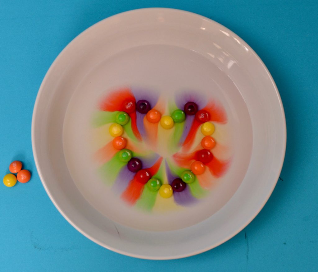 Skittles on a plate in a heart shape. Water has been poured over the skittles and the colour from the sweet has spread through the water.
