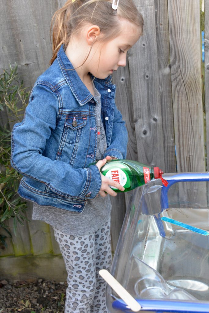 child dropping washing up liquid behind a lolly stick in a container of water as part of a surface tension activity