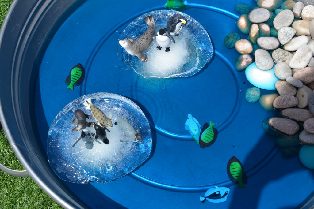 Silver tray with blue water, stones and pretend ice bergs made from lumps of ice. Toy penguins and seals sit on top of the ice ergs