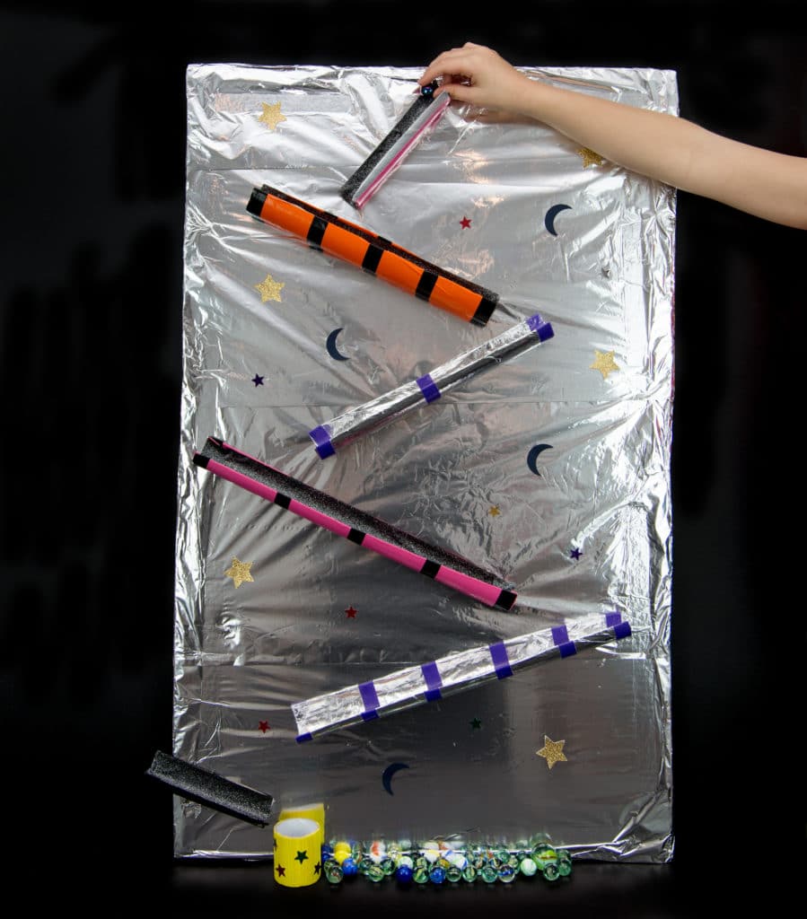 Space Marble Run made by covering a large sheet of cardboard with foil and taping cardboard tubes to it.