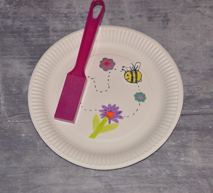 Paper plate with 3 flowers drawn in the centre with a felt tip pen. A dotted line links the flowers. On top of the plate is a magnet wand and a cardboard bee with a paperclip attached.