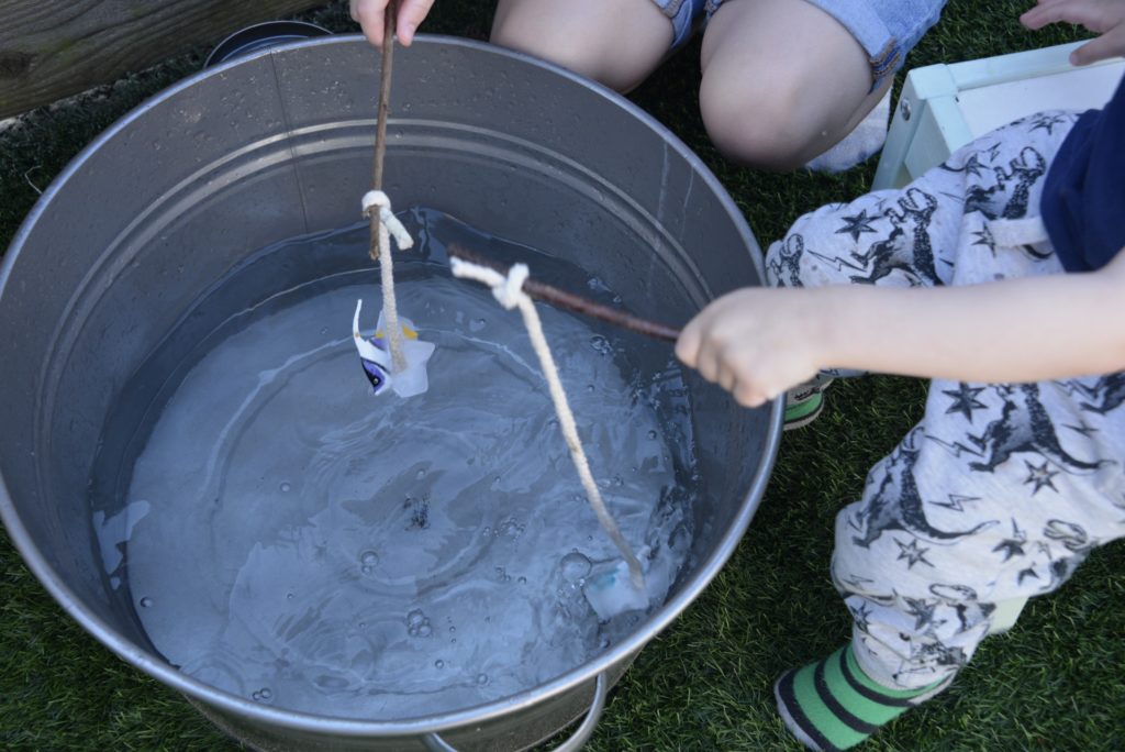 a container of water and two children doing an science experiment where they drop ice cubes into the water like a fishing rod to investigate how fast the ice melts