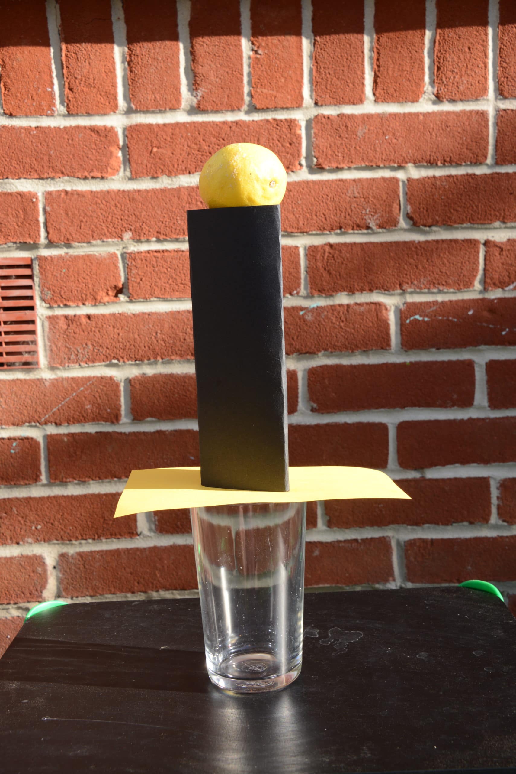A pint glass with a sheet of cardboard on top, with a black cardboard cylinder and a lemon on top