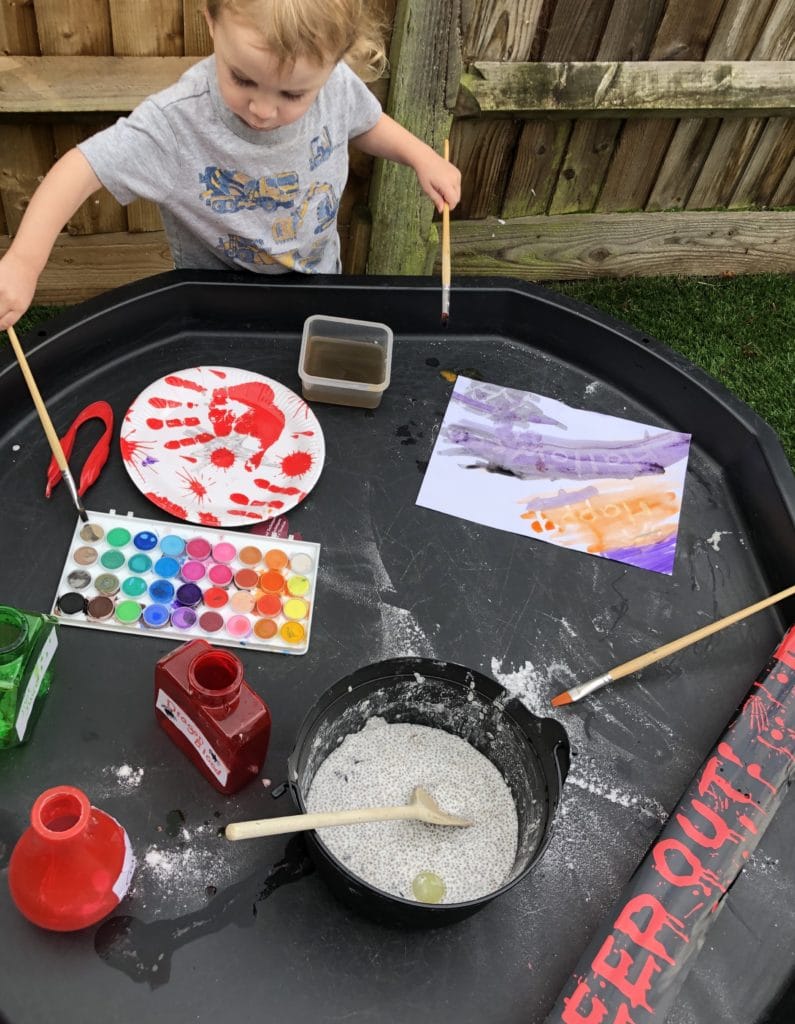 small boy stood next to a tuff tray with Halloween activities set up. Chia seed slime, crayon resist paintings and 