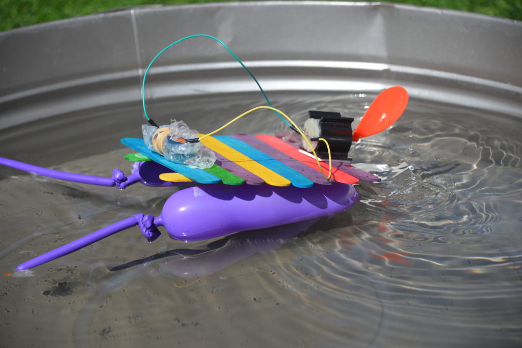 mini motorised boat made with a small motor, AA battery pack covered in bubble wrap to protect it from water and a small propeller. The boat is made from coloured lolly sticks with two long balloons underneath.