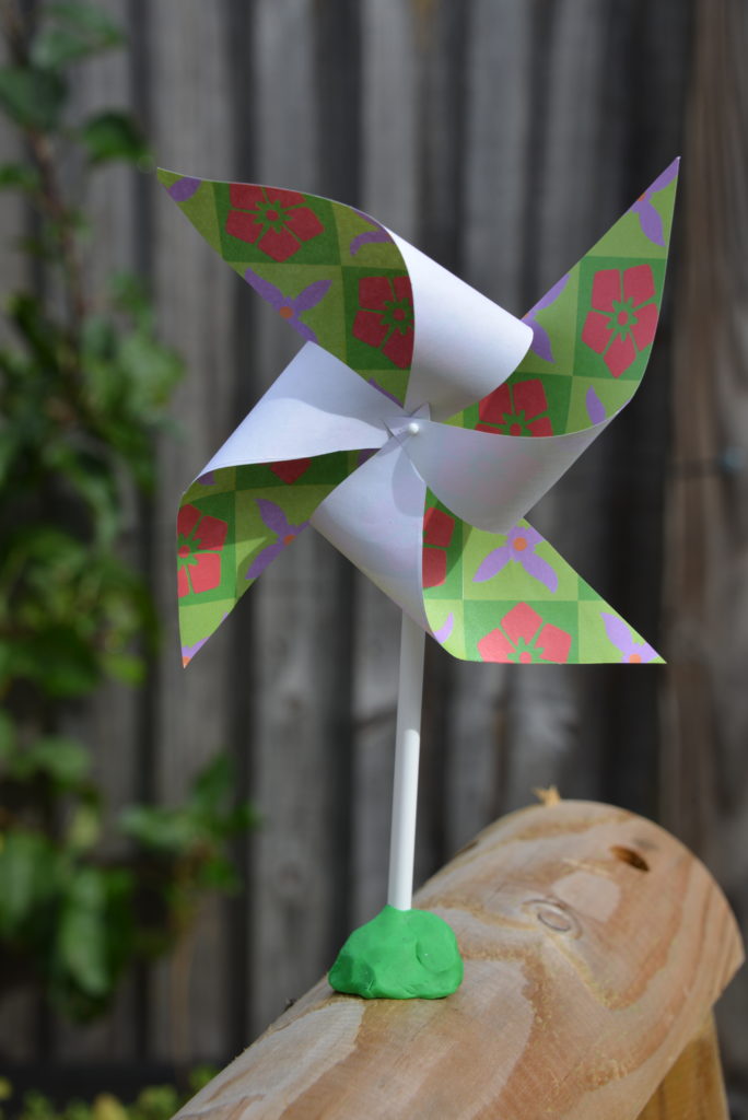 DIY pinwheel made with paper, a pin, pencil and plasticine. Pinwheel is sat outside on a wooden plank.