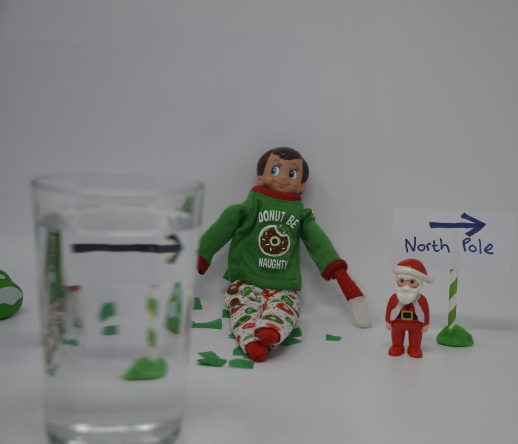 refraction experiment - elf themed. Image shows an elf on the shelf next to a small piece of cardboard with a black arrow on. There is a glass of water in front of the arrow. the arrow is reversed