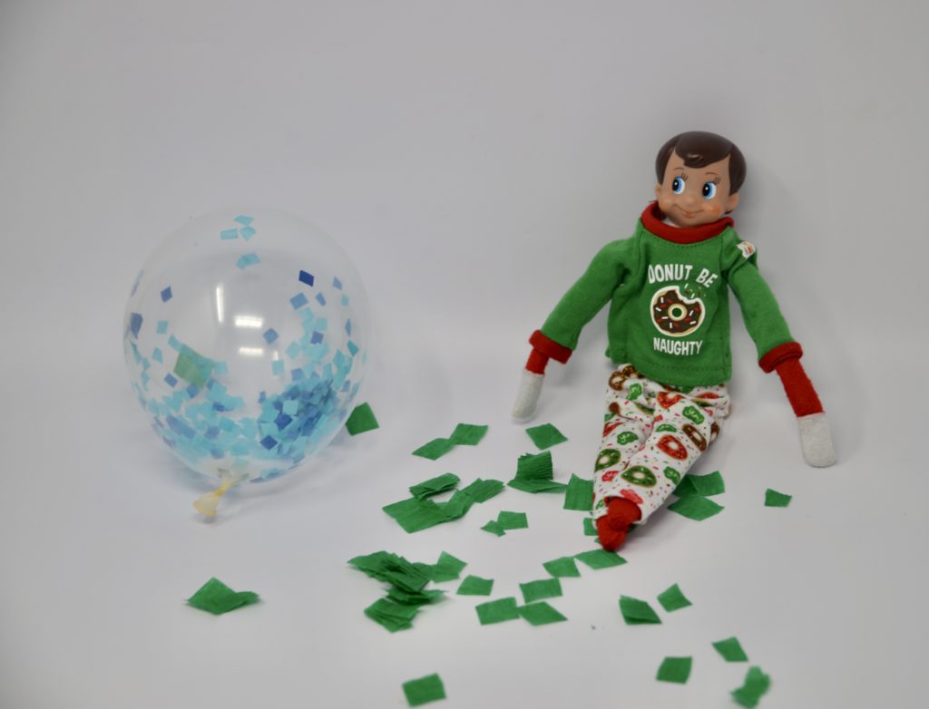 An elf sat near lots of tissue paper pieces in a mess with a balloon. A fun elf trick