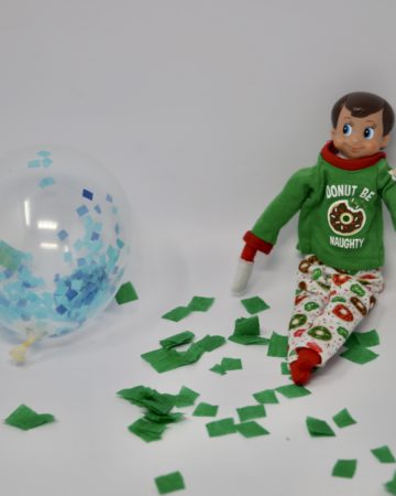 An elf sat near lots of tissue paper pieces in a mess with a balloon. A fun elf trick