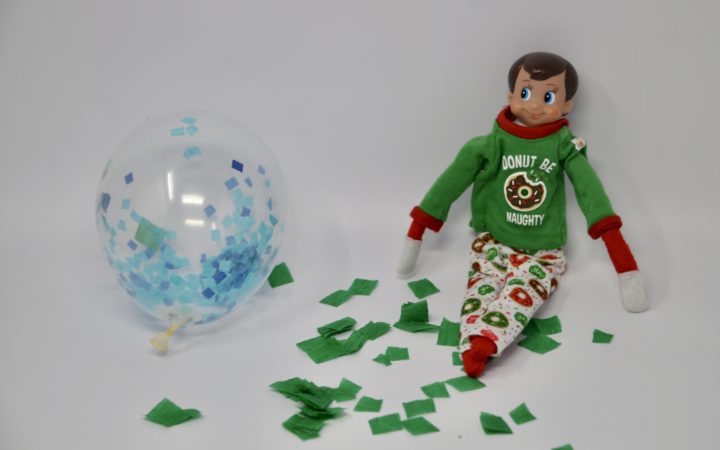 An elf sat near lots of tissue paper pieces in a mess with a balloon. A fun elf trick