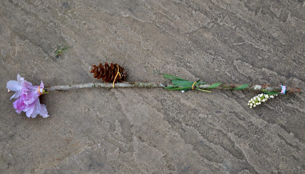 journey stick made from a stick with flowers, a pinecone and other items attached with elastic bands
