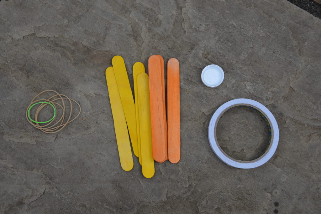 materials needed for a craft stick slingshot - craft sticks, double sided tape, elastic bands and a milk bottle cap