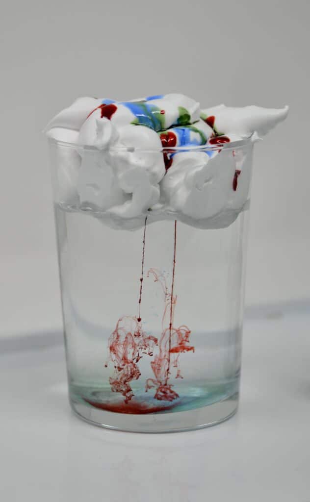 glass full of water with a layer of shaving foam over the top. Food colouring is dripping through the shaving foam to the bottom of the jar.