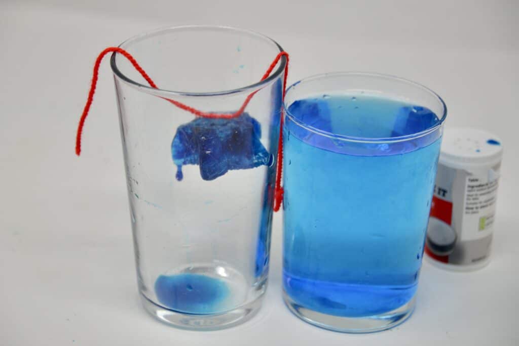 a glass of blue water and an empty glass with a blue ice cube attached to a string suspended over the top