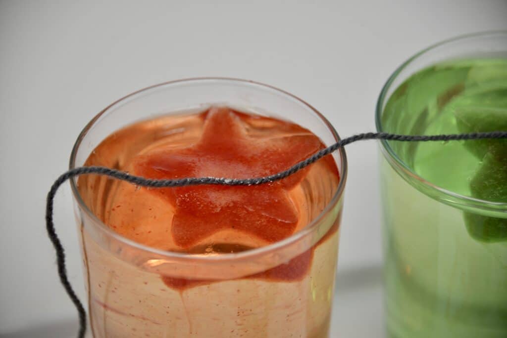 an orange ice cube shaped like a star in a glass of water filled almost to the top with water. A piece or wool sits over the ice cube and salt has been sprinkled on top