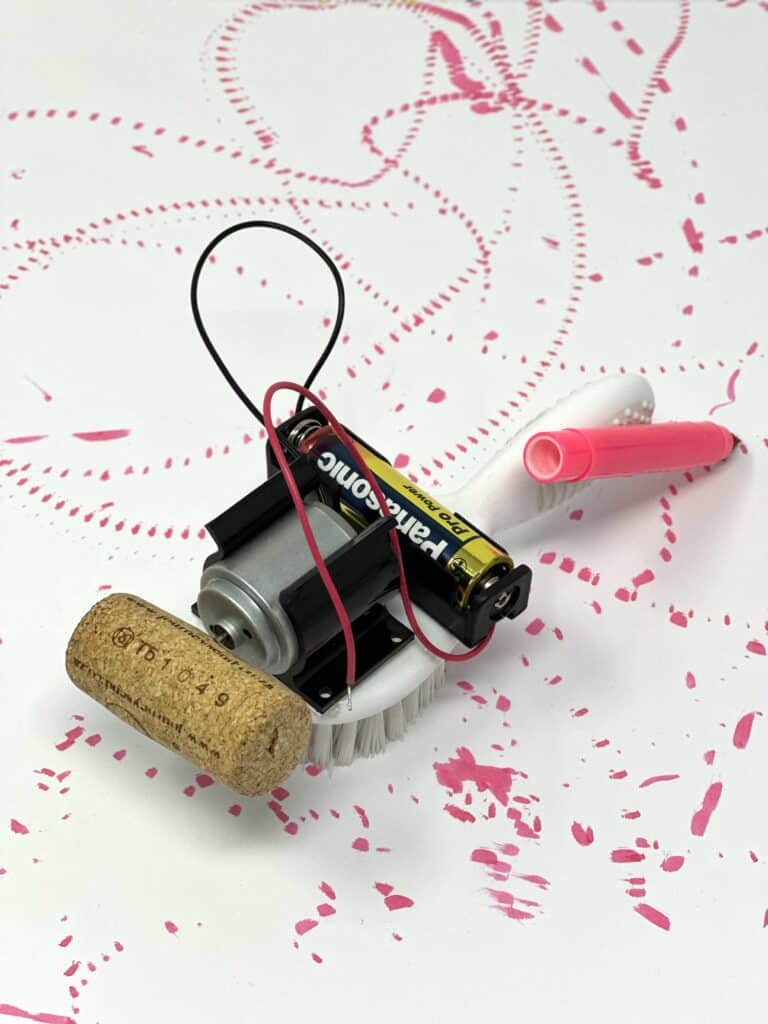 Mini brush bot made with a small white brush. A AA battery pack, motor with a cork and pink felt tip pen are attached to the brush. The robot is sat on a piece of paper with pen marks from the felt tip pen.