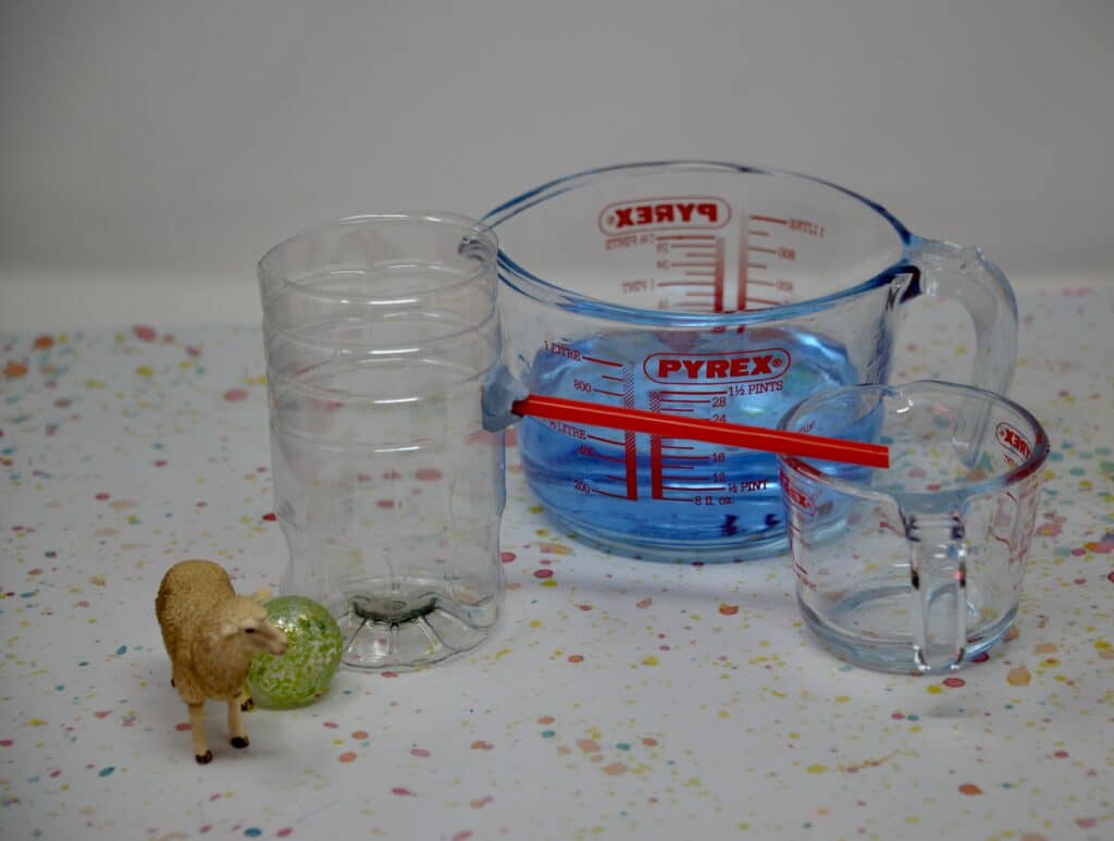 A plastic bottle with a straw attached near the top, and a large jug of blue water. A toy sheep and a marble are next to the bottle