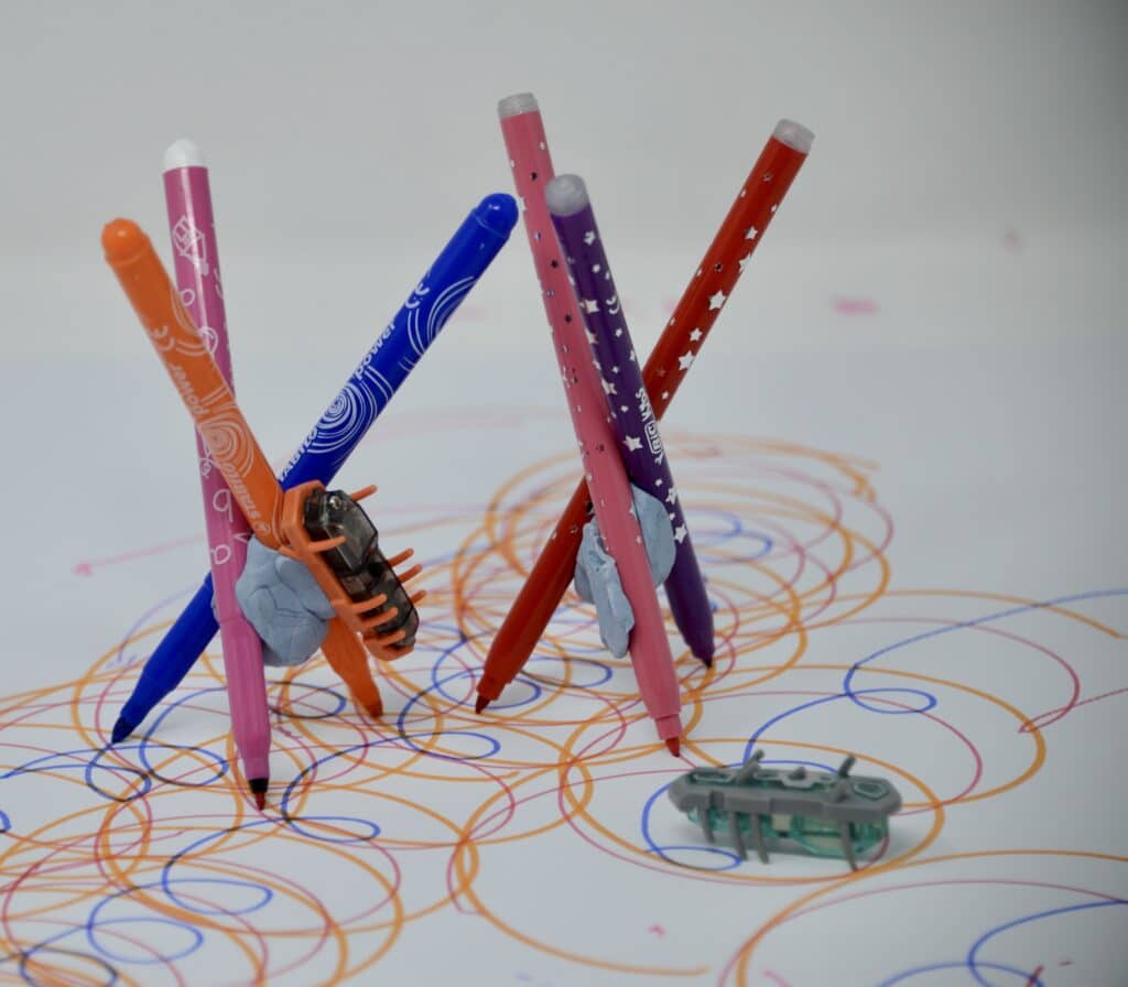 3 felt tip pens held together with blu tack. A Hexbug is attached to one pen and the pens are stood on a large sheet of paper with drawing marks underneath