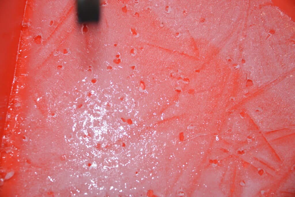 A sheet of ice in a red tray full of holes from where salt has made the ice melt