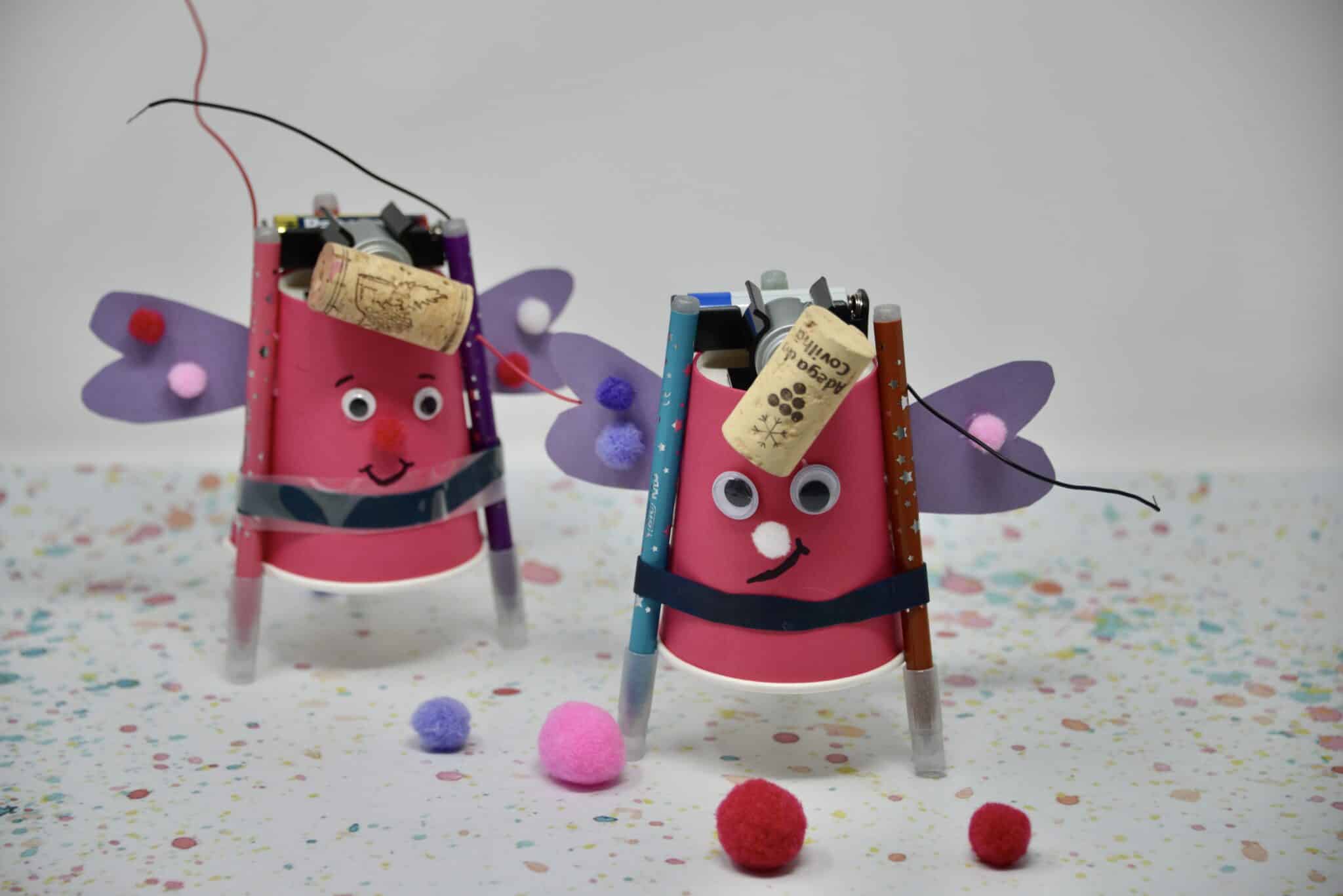 Two pink cups decorated with googly eyes and purple wings to look like love bugs. Theres a motor and AA battery on the top with a cork attached.