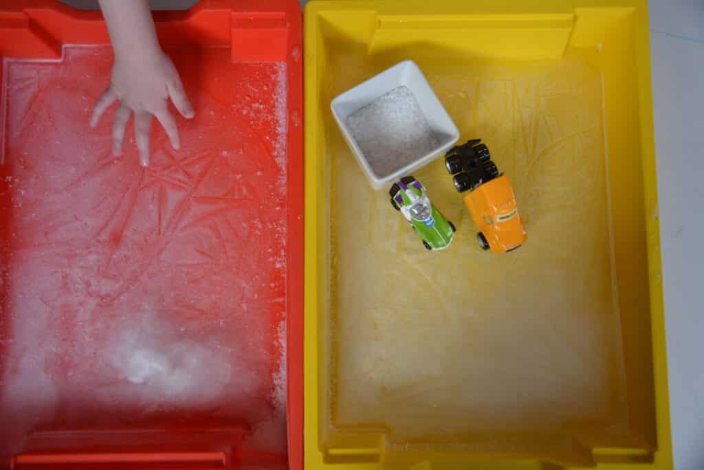 Two trays with ice frozen into the bottom. One tray has a child's hand feeling the ice. The second tray has a small bowl of salt and two push along toy cars on the ice