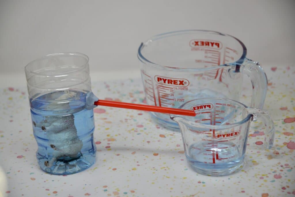 Plastic bottle with the top cut off. A straw is attached to one side near the top and fixed in place with blu tack. The straw hangs over a small jug. The bottle is filled with blue water and a toy sheep is inside.