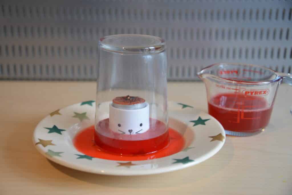 rising water air pressure science experiment - image shows red coloured water on a plate. A candle sits on an egg cup raised out of the water. A pint glass is over the egg cup. The water level has risen higher in the glass than on the plate.
