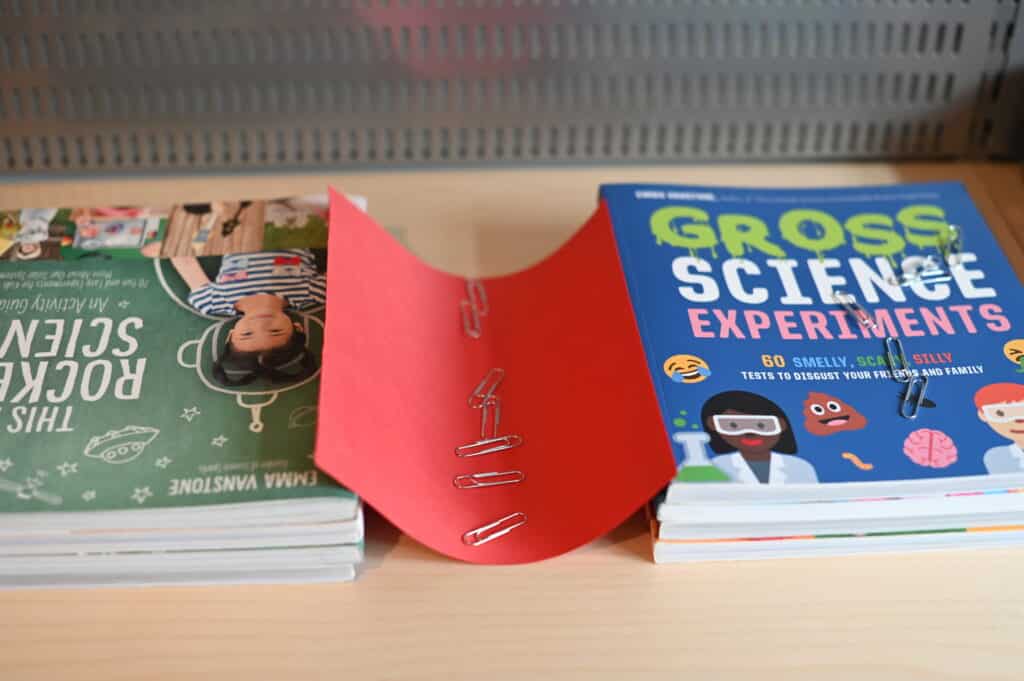 two piles of books with a sheet of paper over the top. Paper clips have been put on the paper bridge which has collapsed