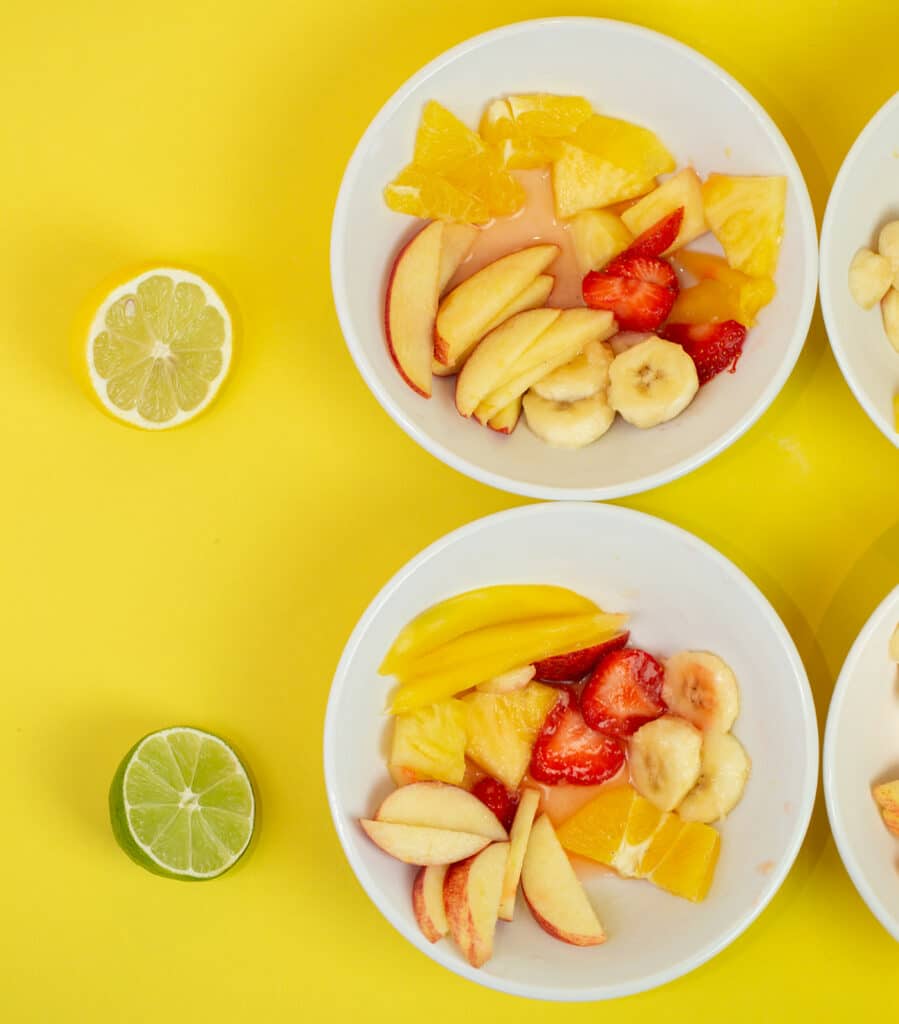 Two bowls of fruit salad. One with half a lemon on the side and one with a lime on the side