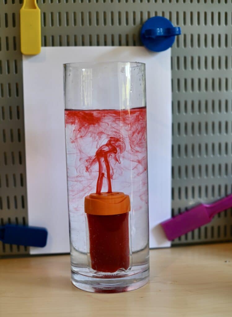 Image shows a large jar filled with cold water. Inside the large jar is a small spice jar filled with hot water coloured red. The red water is escaping upwards through the cold water, demonstrating convection.