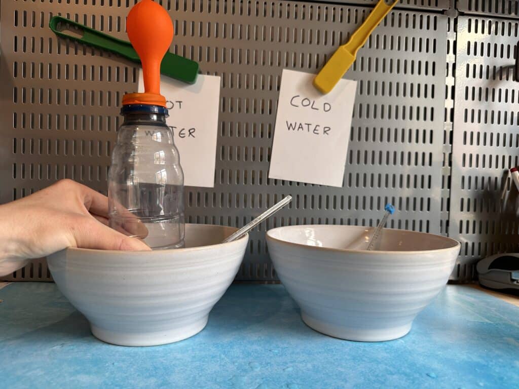 A bowl of hot water and a bowl of cold water for a science experiment. A plastic bottle with a balloon on top is in the hot water bowl. the balloon is slightly inflated.