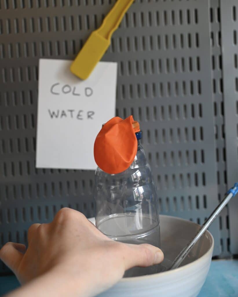 A plastic bottle with a balloon on top in a bowl of cold water. The balloon is deflated